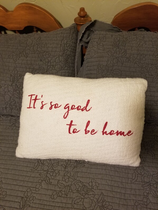 A pillow with the text "It's so good to be home" on a bed with grey bedding and a wooden headboard.