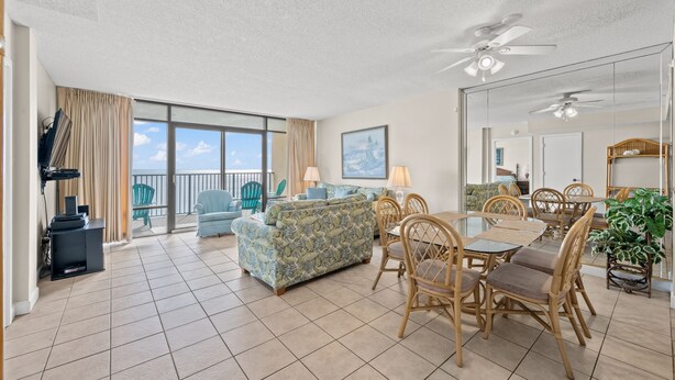 A spacious living area with a large sofa, dining table, and chairs, a balcony with chairs, and a ceiling fan.
