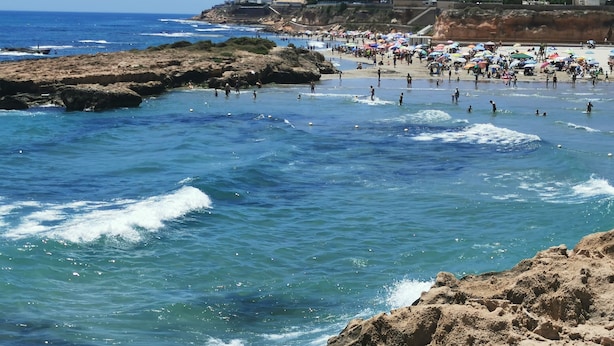 A beach with rocky shores and waves, a crowded beach area with umbrellas, and buildings in the background.