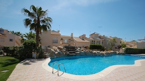 A swimming pool with a stone-tiled deck, surrounded by palm trees and Mediterranean-style buildings.