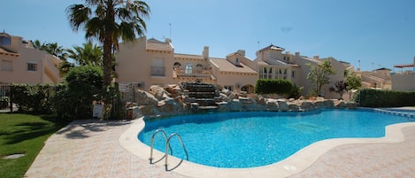 A swimming pool with a stone-tiled deck, surrounded by palm trees and Mediterranean-style buildings.