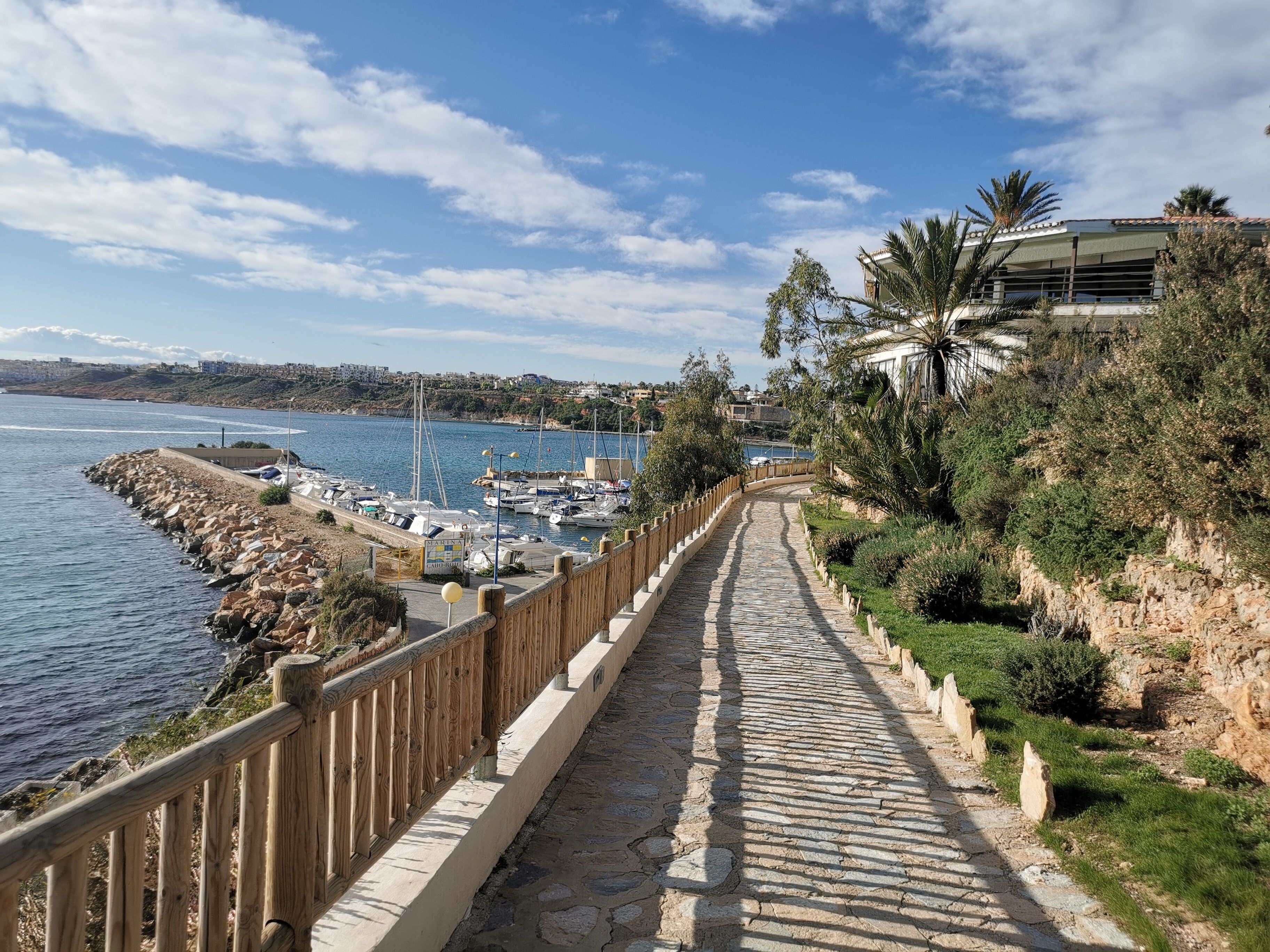 A coastal pathway with a stone pavement, wooden railing, and a view of boats docked at a marina.
