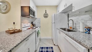A compact kitchen with white cabinets, granite countertops, a microwave, stove, refrigerator, and a hanging kitchen sign. - 2BR Escape | Hot Tub | Parking 4 Cars | Pineville (Charlotte)