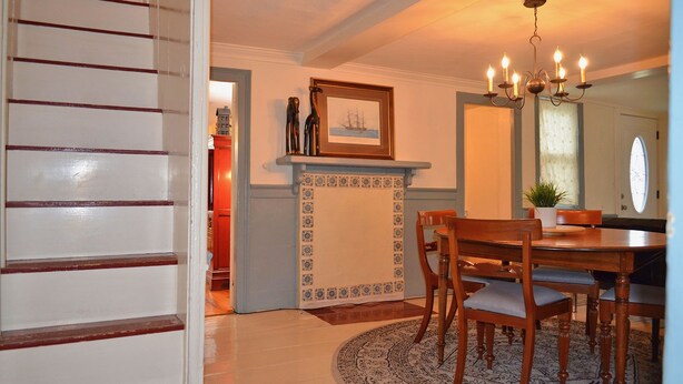 A dining area with a wooden table and chairs, a patterned rug, a chandelier, and a framed picture above the fireplace. - The Captain John Butman House (Rockport)
