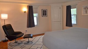 A bedroom with a bed, a chair, a small table, a lamp, and two windows with curtains. - The Captain John Butman House (Rockport)