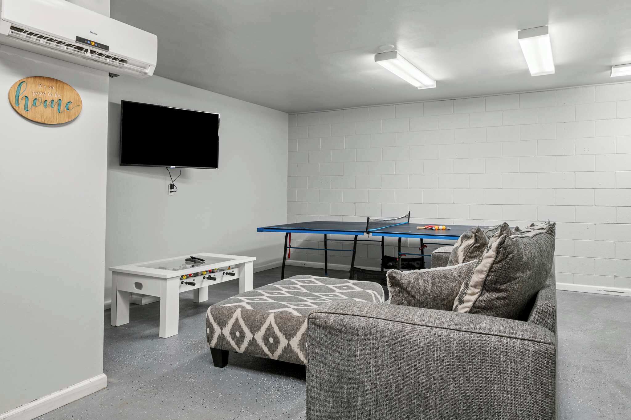 A room with a white brick wall, a flat-screen TV mounted above a small white table, a ping pong table, and a grey patterned sofa.
