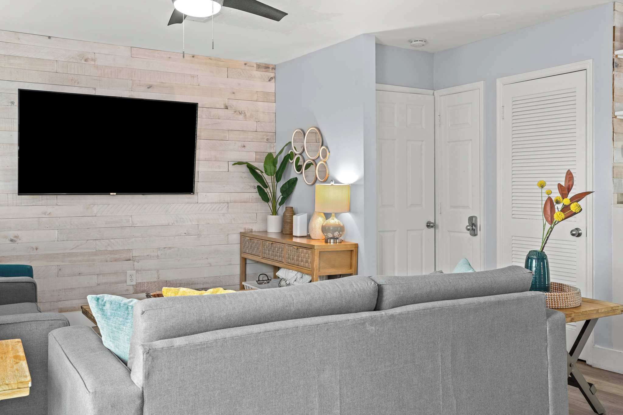 A living room with a flat-screen TV mounted on a wooden wall, a grey sofa, a wooden side table with a lamp, and a plant.