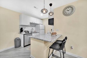 A compact kitchen with a granite countertop, white cabinets, a stainless steel refrigerator, and a black trash bin.