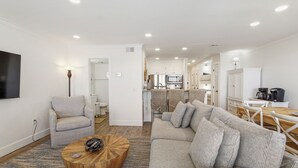 A modern living room with a sofa, armchair, and coffee table. A kitchen is visible in the background with a dining area.
