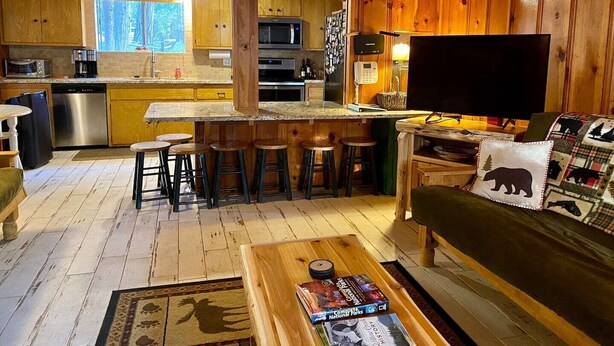 A cozy cabin interior with a wooden interior, a kitchen area, a dining space, and a living room with a TV and books. - Bear’s Lair Cabin, Willow Cove, Bass Lake (Bass Lake)