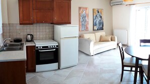 Cuina compacta amb armaris de fusta, fogó i nevera. Hi ha una zona de dinar amb taula i cadires, i un sofà. Dues pintures abstractes adornen la paret.