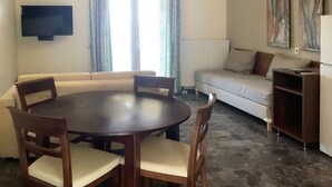 A living room with a round wooden table, four chairs, a sofa, and a television mounted on the wall. - 03 | Sea View - One Bedroom Apartment (Palaiokastritsa)