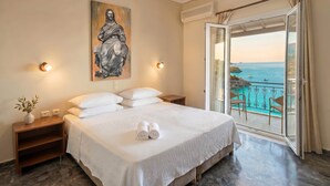 A bedroom with a bed, a painting, bedside lamps, a nightstand, and a view of the sea through a balcony. - 03 | Sea View - One Bedroom Apartment (Palaiokastritsa)