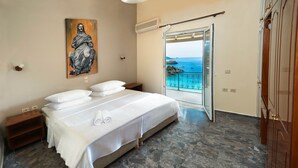 A bedroom with a bed, a painting, a nightstand, a wardrobe, and a view of the sea through a glass door. - 03 | Sea View - One Bedroom Apartment (Palaiokastritsa)