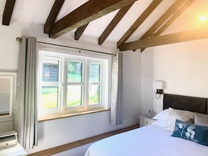 Ein Schlafzimmer mit einem großen Bett, einem Nachttisch, einem Beistelltisch, einem Fenster mit Vorhängen und einem Holzbalken an der Decke.