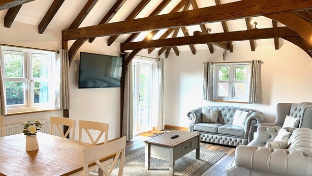 A cozy living room with a wooden ceiling, a grey sofa, a white armchair, a wooden dining table, and a TV mounted on the wall. - Flint Cottage - Sleeps 6 - Private Woodland (Hailsham)