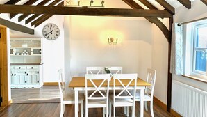A dining area with a wooden table and white chairs, a clock on the wall, and a kitchen in the background. - Flint Cottage - Sleeps 6 - Private Woodland (Hailsham)