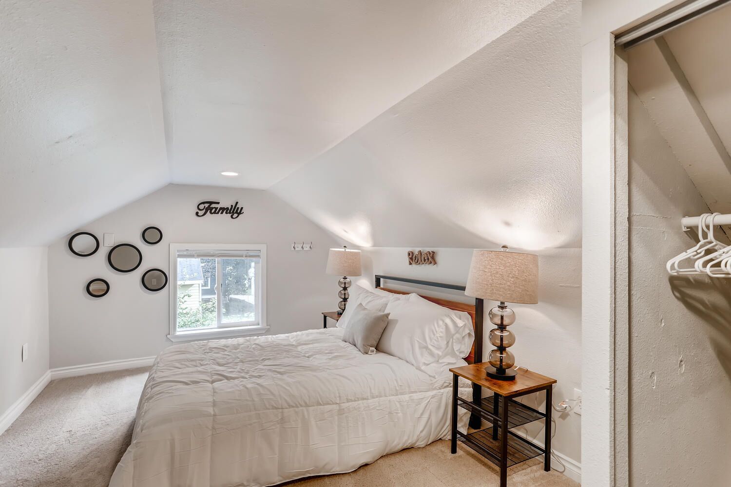 Ein Schlafzimmer mit schrägen Wänden, einem Bett mit weißer Bettwäsche, einem Nachttisch mit Lampe, einem Fenster und Wanddekorationen, darunter ein Schild mit der Aufschrift 'Family' und ein Schild mit 'Rolex'.