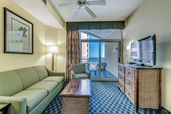 A hotel room with a sofa, a small table, a television, a ceiling fan, a balcony with a view, and a framed picture on the wall.