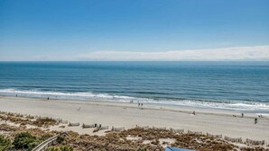 A beach with a clear sky, calm sea, and a sandy shore with a few people. - · NEW! Stunning Oceanfront Condo -Amazing Water Amenities! (Myrtle Beach)