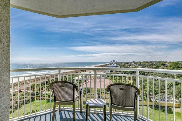 A balcony with two wicker chairs and a small table, overlooking a beach and distant city skyline.