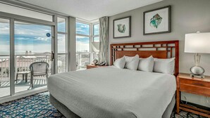 A hotel room with a large bed, a nightstand, a lamp, a chair, and a view of the city through large windows. - · 3Bdrm Deluxe Oceanfront Condo - Gorgeous Pools - Top Rated Resort! (Myrtle Beach)