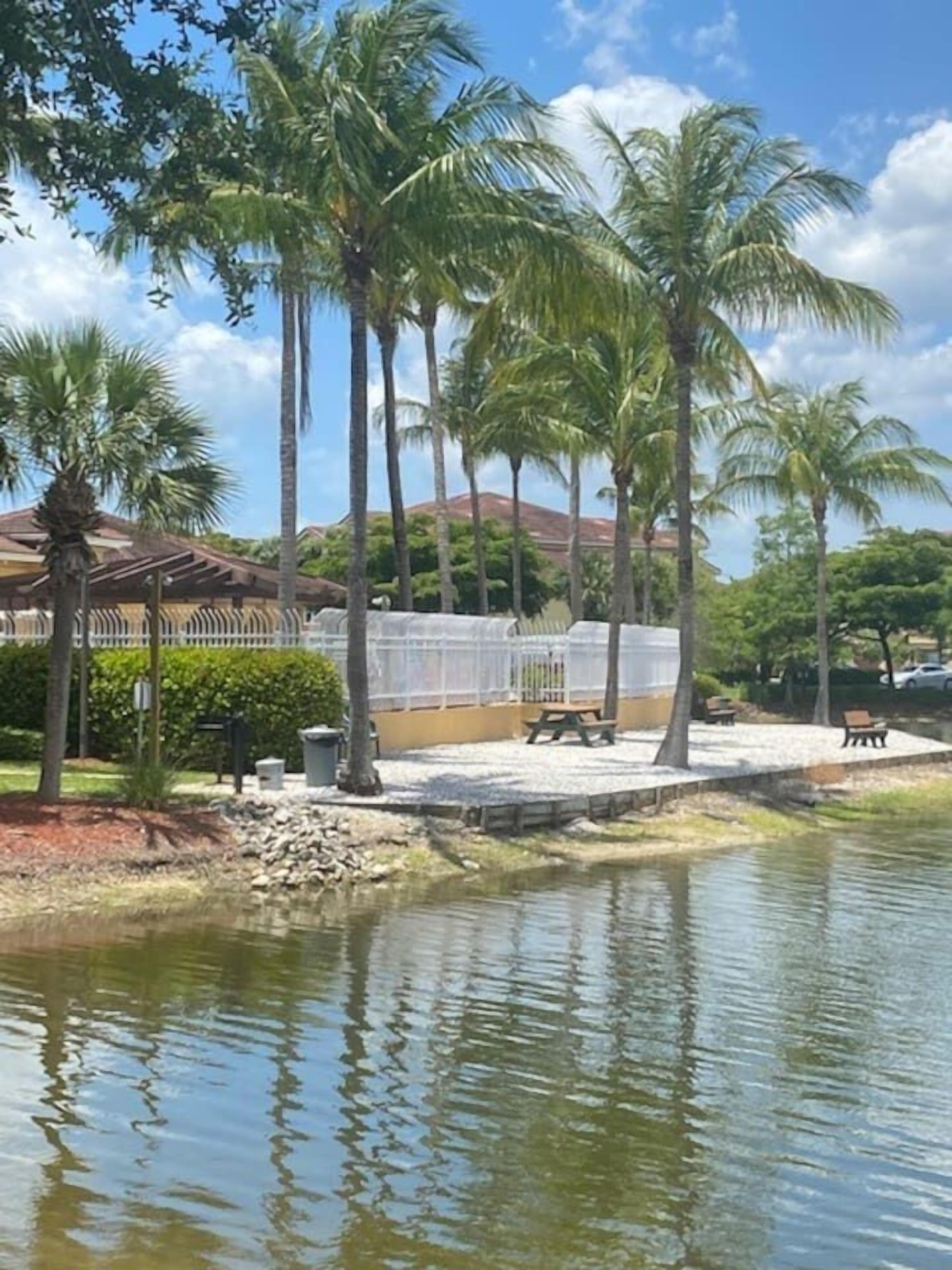 A serene lakeside setting with palm trees, a gazebo, and a fenced area with benches.