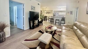 Ein Wohnzimmer mit einer Couch, einem Couchtisch und einem Fernseher, ein Essbereich mit Stühlen und eine Küche im Hintergrund.