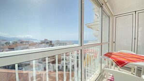 A balcony with a view of the sea and buildings, a table with a patterned tablecloth, and a chair with a pink cushion.