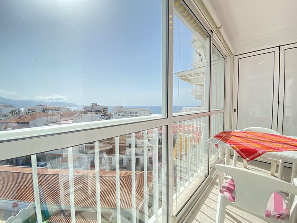 Ein Balkon mit Blick auf das Meer und Gebäude, ein Tisch mit gemustertem Tischläufer und ein Stuhl mit rosa Kissen.