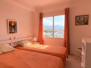 Ein Schlafzimmer mit zwei Betten, einem Fenster mit Vorhängen, einem Nachttisch mit Lampe und einem Bild an der Wand.