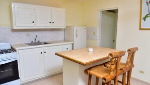 Een compacte keuken met witte kastjes, een gasfornuis, een spoelbak en een klein eiland met een tafel en twee rieten stoelen.
