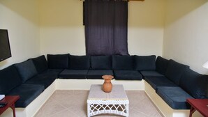 Een woonkamer met een hoekbank, een kleine tafel met een vaas, en een televisie aan de muur.