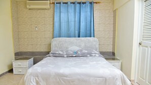 Een slaapkamer met een bed, nachtkastje, dressoir, een raam met blauwe gordijnen en een airconditioning.