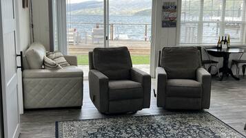 Een woonkamer met uitzicht op een meer, twee grijze fauteuils, een witte bank en een kamerkleed met een patroon.