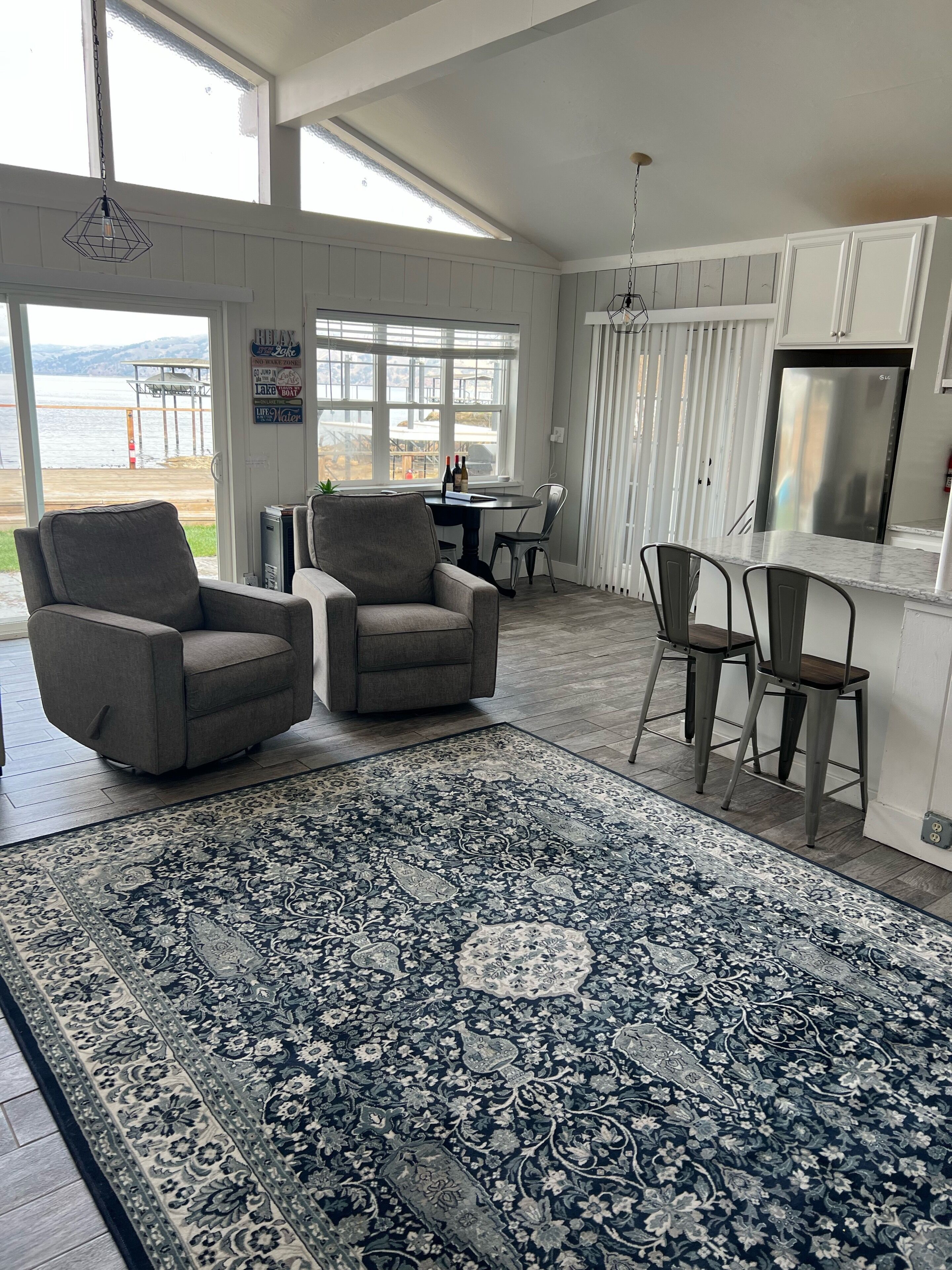Un vaste espace de vie agrémenté d’un grand tapis, de deux fauteuils, d’une table à manger avec des chaises, et offrant une vue sur un lac grâce à de grandes fenêtres.