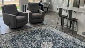 Amplia sala de estar con un tapete grande, dos sillones, una mesa de comedor con sillas y vistas a un lago a través de amplias ventanas.