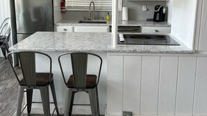 Una cocina compacta con armarios blancos, un refrigerador de acero inoxidable, una encimera de mármol, dos taburetes de bar, una lámpara colgante y una decoración con la bandera estadounidense.