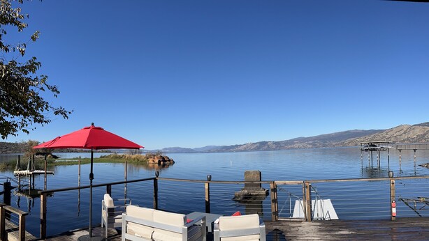 Una terraza de madera con un paraguas rojo, sillas de lounge blancas y vistas a un lago tranquilo con montañas al fondo.