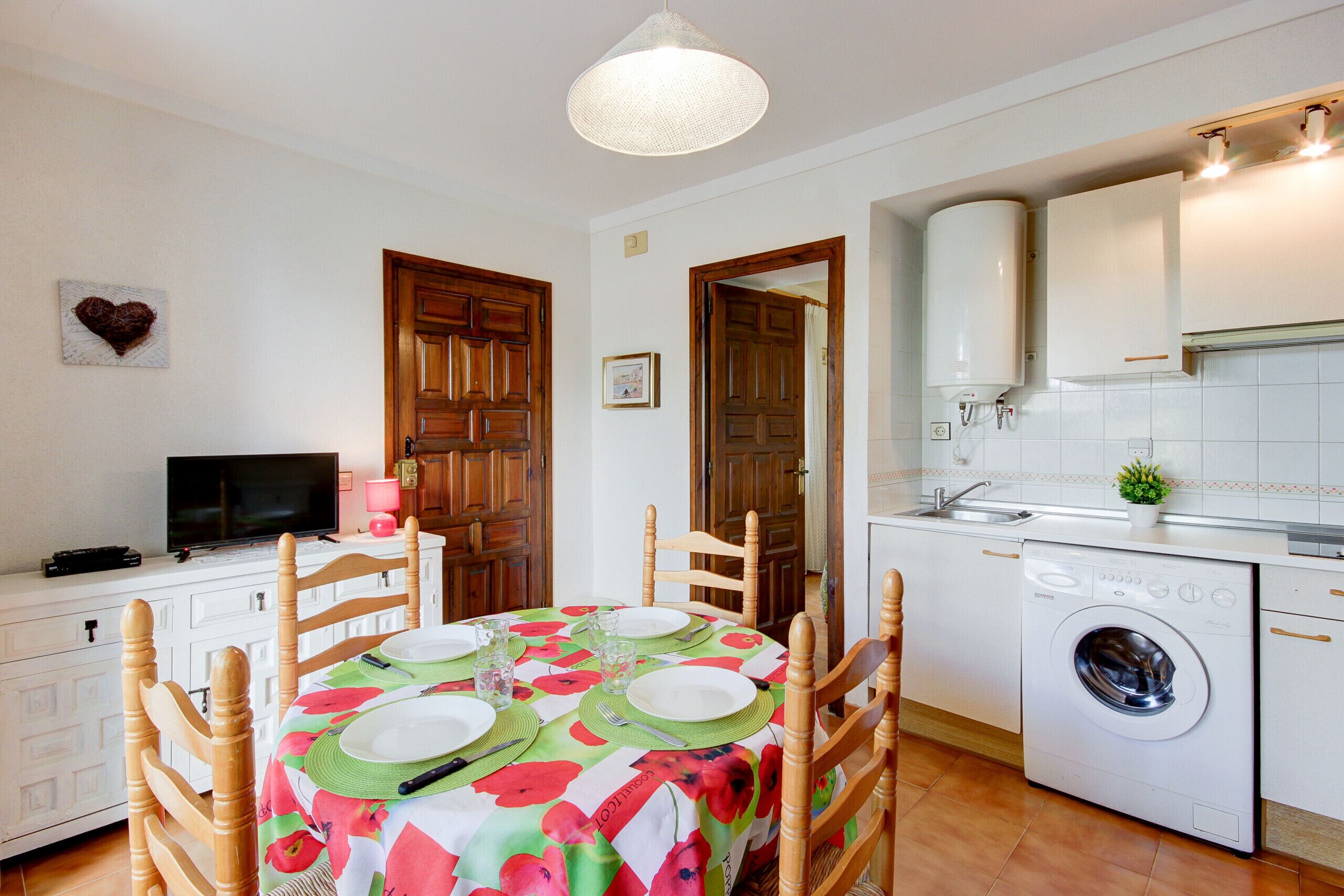 A compact kitchen with a washing machine, wooden cabinets, and a dining area with a table set for two.