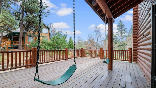 Une terrasse en bois avec une balançoire, un espace abrité et une vue sur un chalet et des arbres.