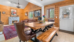 L’intérieur d’une cabane en rondins, avec une table à manger en bois, des fauteuils en cuir, une cheminée en pierre et une grande fenêtre équipée de stores.