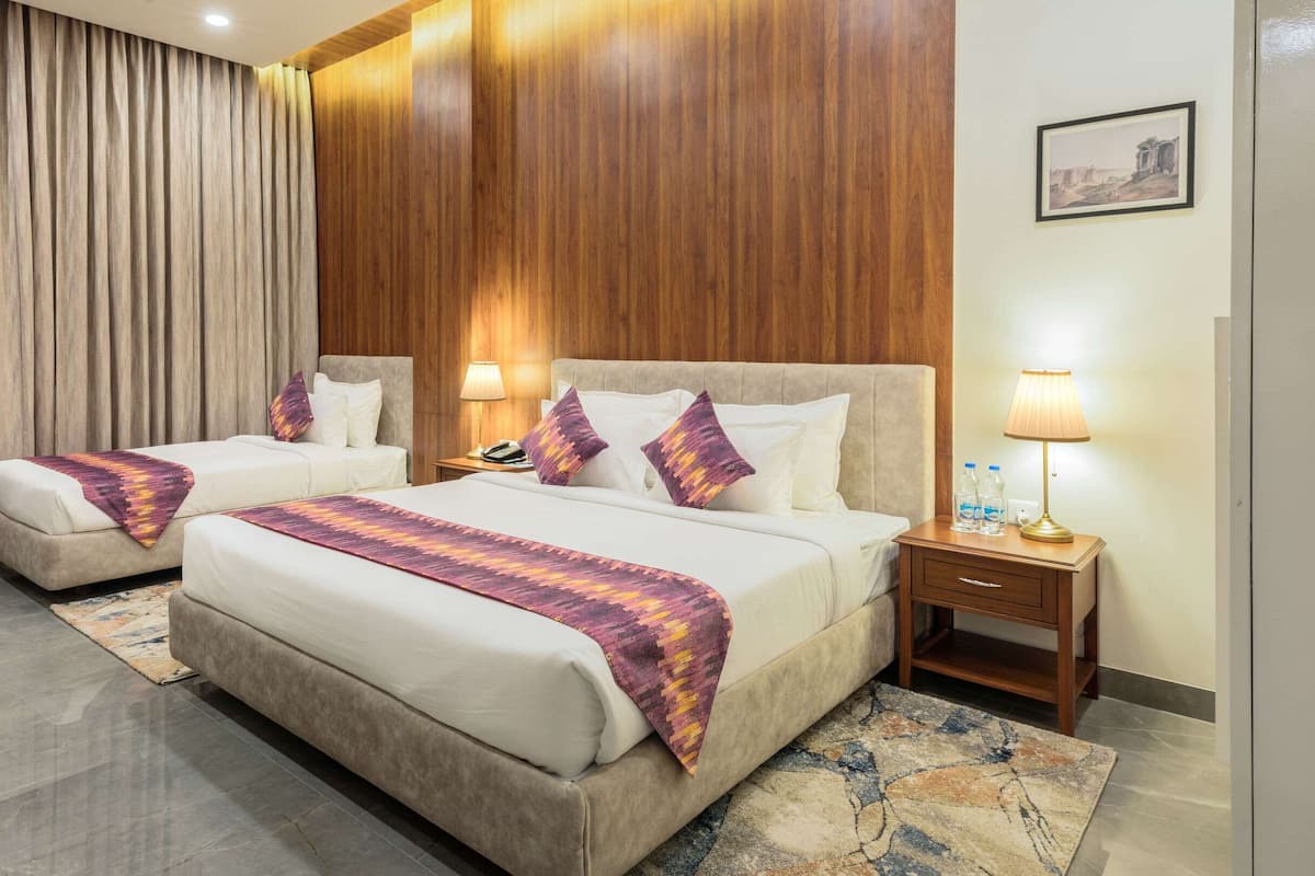 A hotel room with two beds, a wooden panel wall, a large window with curtains, a nightstand with a lamp, and a painting on the wall.