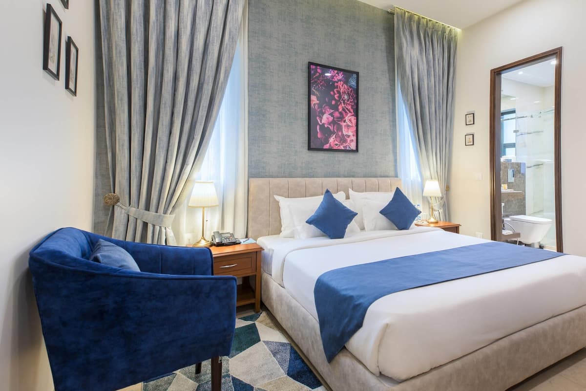 A hotel room with a large bed, two bedside tables, a desk, a chair, and a bathroom visible through a glass door.