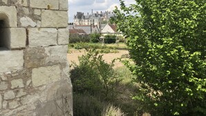 A stone wall with a small opening, a green bush, and a view of a large building with multiple roofs in the background.