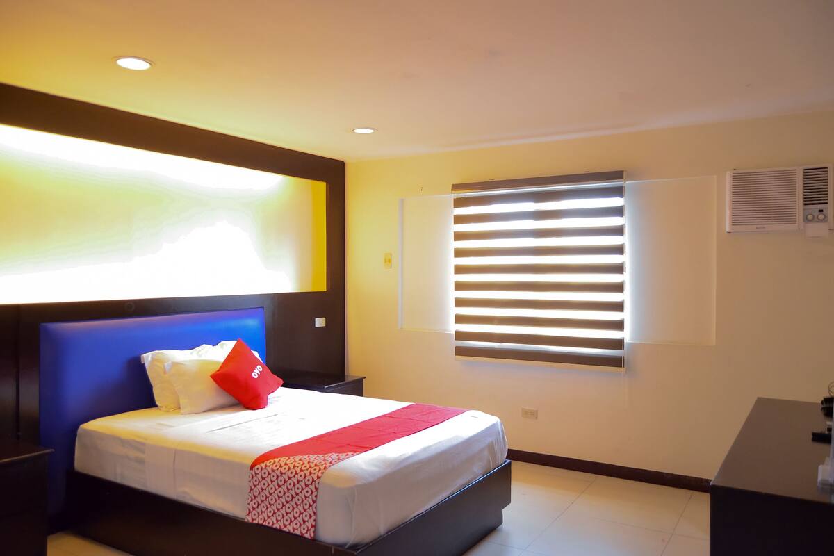 A hotel room with a bed, a nightstand, a desk, and a window with blinds.