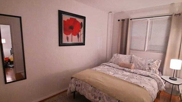 A bedroom with a bed, a mirror, a framed picture, a bedside table with a lamp, and a window with curtains.