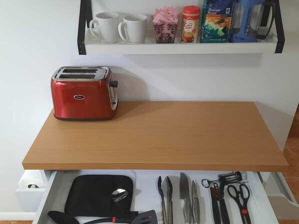 Una kitchenette con una tostadora roja sobre un estante de madera, una cafetera y una taza de café en la encimera. Abajo, un cajón contiene una billetera negra, cubiertos y un par de tijeras.