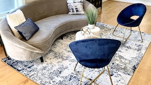Een moderne woonkamer met een gebogen grijze bank, twee blauwe fauteuils, een kamerbreed tapijt met een patroon en een kleine witte tafel met een plant.