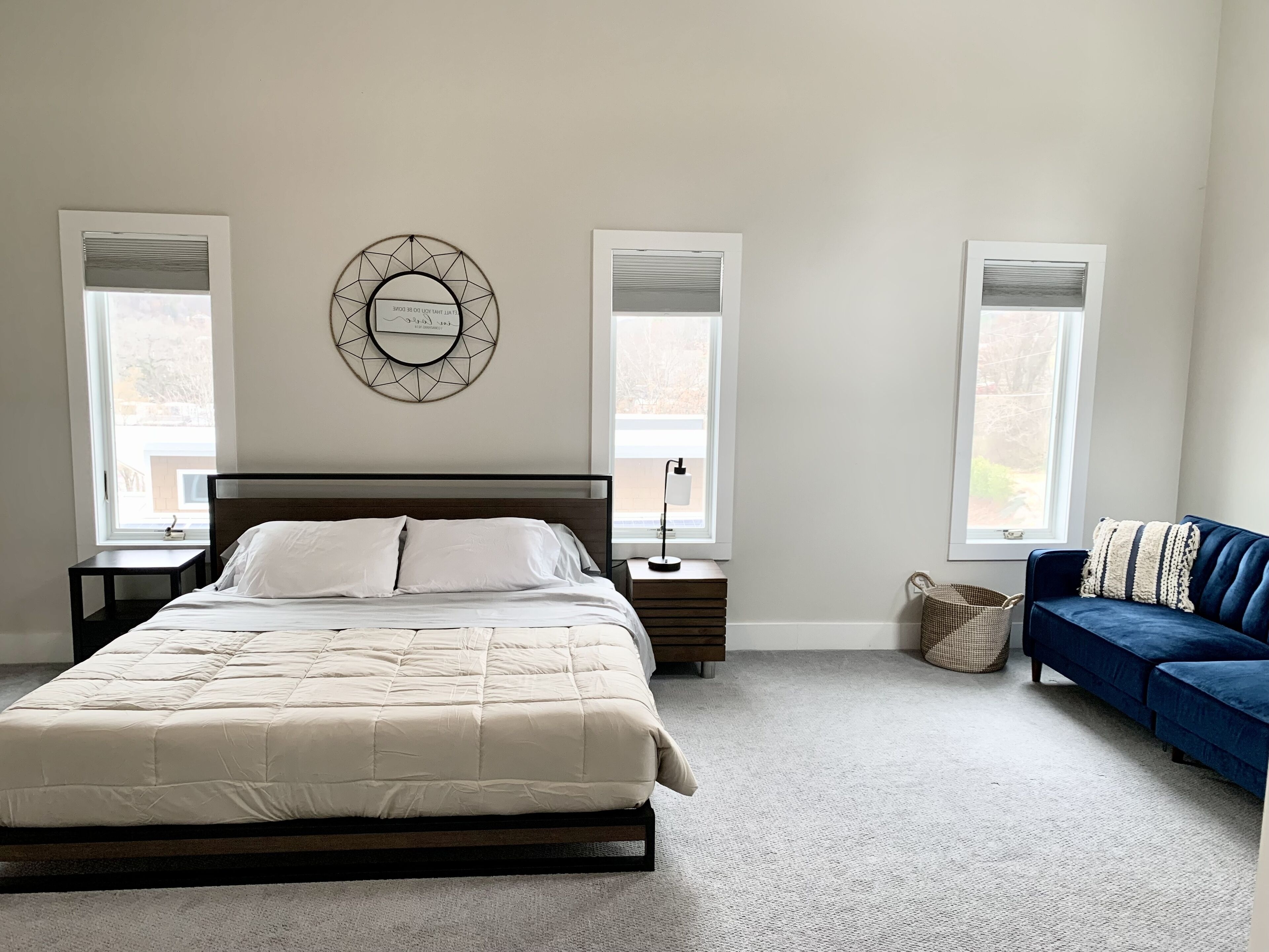 A bedroom with a bed, two bedside tables, a wall clock, a blue sofa, and two windows.
